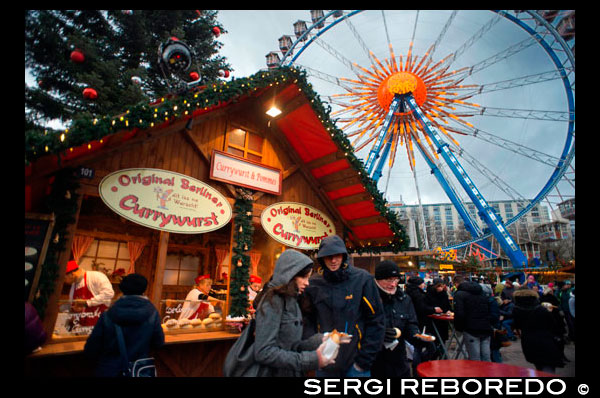 Rueda de la fortuna y currywurst en el mercado de Navidad en frente de la fuente Neptunbrunnen , Alexanderplatz , Berlín . Snack-bar en Alexanderplatz . Las campanas , guirnaldas , villancicos, árboles decorados ... Navidad ha llegado definitivamente . Este momento especial del año, comienza a hacerse sentir en todos los rincones de la capital. No hay muchas ciudades en el mundo que saben cómo dar la bienvenida a la Navidad como Berlín. Es un hecho bien conocido de que el pueblo alemán encanta celebrar la Navidad. Berlín no quiere ser eclipsado por ciudades como Munich o Dresden , que son más ampliamente conocidos por sus celebraciones de Navidad. Cuenta con mercados de Navidad sesenta repartidas por toda la ciudad : tradicional o alternativa , especializada en artesanías o alimentos , gratuito o de pago , hay un mercado para todos los gustos . Uno de los mercados más turísticas y comerciales , pero al mismo tiempo uno de los más característicos , se lleva a cabo en los alrededores de la plaza Alexanderplatz . Entrando por la Karl- Liebknecht- Strasse , a lo largo de la plaza y terminando por Alexa , se encuentra uno de los mejores Weihnachtsmarkt la ciudad tiene para ofrecer. Una vez pasado la puerta principal brillante empezamos a sentir el verdadero espíritu de la Navidad . La canción " Feliz Navidad a todos " y otros clásicos se pueden oír justo al lado de la estatua Neptunbrunnen , que a su vez está rodeado por una de las muchas pistas de patinaje sobre hielo que adornarán la ciudad este mes. Desde el cielo, el timón de transbordador de 50 metros mira hacia usted. Aquellos que desafían las alturas podrán disfrutar de hermosas vistas de todo el mercado y también el distrito Mitte de los alrededores. Hay callejones largos en los que las cabinas están colocados en filas , las pequeñas casas de madera , techos con nieve artificial , música, campanas , trineos y luces por todas partes. Es sin duda una de las maravillas del invierno.
