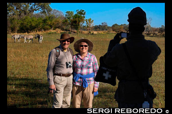 Una parella de turistes es fotografia amb les zebres de fons en el safari a peu realitzat en els voltants del campament Eagle Island Camp d'Orient Express, als afores de la Reserva d'Animals de Moremi, a Botswana. Després atalaiar l'horitzó, Botsualo, el nostre guia, ens autoritza a baixar per instal · lar les tendes de campanya i iniciar una caminada al capvespre. "No heu de preocupar-vos, en aquesta zona del delta no hi ha depredadors" ens diu quan veu les nostres cares, entre espantades i il · lusionades per caminar per aquesta zona. Normalment als parcs naturals no es pot caminar però en zones determinades, amb guia si. Aquesta és una. Sigilosos avancem reconeixent enormes petjades d'elefants i excrements de micos, fins a una zona de pastures on diversos grups de zebres, nyus i antílops pasturen al seu aire. Descobrim un esquelet de girafa. Més lluny, al costat d'un enorme baobab, un elefant solitari. No podem acostar-nos massa però el ser-hi, sense la protecció del cotxe, en plena natura, en el seu terreny, és una sensació meravellosa i alliberadora a parts iguals. A mesura que cau el sol ens retirem. Encara que sigui la millor hora per observar els animals hem de tornar al campament, a encendre el foc i cuinar el sopar. No hem fet cap esforç, però la naturalesa en estat pur sembla que fa entrar la gana. Després del sopar, la humitat de la nit porta el fred als nostres ossos encara que per sort tenim les brases per escalfar-nos. En el silenci gairebé total qualsevol soroll és perceptible, així que ens vam quedar muts en sentir, a només uns metres de distància, soroll d'aigua, petjades en fang, branques trencant, arbres sacsejats ... "No us mogueu ni encengueu cap llum", ens va dir Botsualo deixant-nos sols mentre sortia a mirar, sigil · lós. Vam deduir que un soroll així solament podia ser un elefant. A mesura que s'allunyava el soroll el guia va tornar amb un enorme pal a la mà. "Els elefants són els animals més perillosos. Només dormen 3 o 4 hores i segueixen passejant-se per la nit. I poden no veure la botiga de campanya i ... "ens va dir, deixant la frase inacabada. "Una vegada", ha prosseguit, "per espantar a un vaig haver colpejar perquè s'havia ficat, molt molest, en un campament, on teníem torxes". Així és, al delta, dormint en algun illot, no hi ha tanques ni proteccions. És pura naturalesa, el seu territori, aquesta és la bellesa. I una cosa així no té preu.