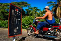 Atiu Island. Cook Island. Polynesia. South Pacific Ocean. An inhabitant of Atiu driving with his bike on the roads of the island next to a poster from Keep Left. Although Atiu is great for walking, a scooter (motorbike) or bicycle will allow you to explore more of the island at your own pace. Atiu Villas rents out scooters, bicycles and a jeep. Their own guests have priority but they will also rent to others. There are other businesses on the island renting out scooters though too and your accommodators can help organize this. You should let them know you might want to rent a scooter when booking your accommodation. Atiu Guesthouse and Atiu Homestay also have a four-wheel drive vehicle they rent out. Again it would pay to make enquiries and book ahead to ensure it is available. If you are doing some tours (very highly recommended) and some walking, you may only want to hire a scooter or jeep for one day - depending of course on how long you are staying on Atiu. Because of the small and personalized nature of tourism on Atiu, all of the accommodators will collect you from the airport and bring you to your accommodation. Transport may be on the back of a ute - travel like a local and enjoy the view! Fuel on Atiu is expensive and there is usually a cost for airport transfers although it may be included in your accommodation package. Check beforehand to avoid any misunderstandings when it's time to pay the bill. There is no public transport or taxi service on Atiu.