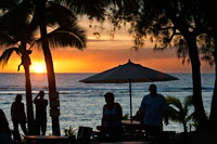 Rarotonga Island. Cook Island. Polynesia. South Pacific Ocean. Sunset on the beach in Hotel Crown Beach Resort & Spa.  Lit. Tourists taking photos. Parasols.  The Crown Beach Resort comprises of 22 villas nestled in four and a half acres of tropical garden, which leads onto a beautiful stretch of white sand beach. The stunning swimming pool has fantastic views of the beach and the serene flame tree and gardens; an ideal location for relaxing with a good book. After a hard days sunbathing or snorkelling you can make your way to the on-site spa for a therapeutic massage or treatment. Along with complimentary tropical breakfast and afternoon tea, this resort is home to two restaurants: The Windjammer, which serves delicious fish of the day and home made bread, and the beach side Cabana Bar and Grill, for more casual dining. Each comfortable villa is self contained ensuring maximum comfort, allowing you to enjoy natural warmth and charm of the Cook Islands in this attentive resort.