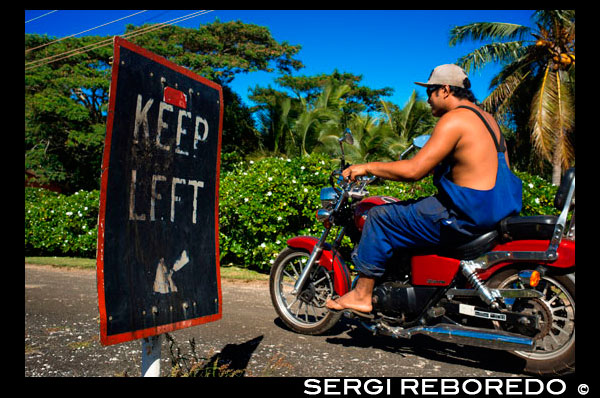 Atiu Island. Cook Island. Polynesia. South Pacific Ocean. An inhabitant of Atiu driving with his bike on the roads of the island next to a poster from Keep Left. Although Atiu is great for walking, a scooter (motorbike) or bicycle will allow you to explore more of the island at your own pace. Atiu Villas rents out scooters, bicycles and a jeep. Their own guests have priority but they will also rent to others. There are other businesses on the island renting out scooters though too and your accommodators can help organize this. You should let them know you might want to rent a scooter when booking your accommodation. Atiu Guesthouse and Atiu Homestay also have a four-wheel drive vehicle they rent out. Again it would pay to make enquiries and book ahead to ensure it is available. If you are doing some tours (very highly recommended) and some walking, you may only want to hire a scooter or jeep for one day - depending of course on how long you are staying on Atiu. Because of the small and personalized nature of tourism on Atiu, all of the accommodators will collect you from the airport and bring you to your accommodation. Transport may be on the back of a ute - travel like a local and enjoy the view! Fuel on Atiu is expensive and there is usually a cost for airport transfers although it may be included in your accommodation package. Check beforehand to avoid any misunderstandings when it's time to pay the bill. There is no public transport or taxi service on Atiu. 