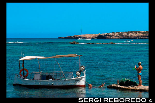 Els Pujols beach in Formentera. Tourists taken pictures with traditional fishing boat in summer day. Llaüt. Magnificent beach in the tourist area of Es Pujols, where you can find all the services: accommodation, shopping, restaurants, water sports.