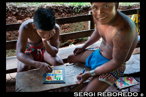 Playing Parcheesi in the village of the Native Indian Embera Tribe, Embera Village, Panama. Panama Embera people Indian Village Indigenous Indio indios natives Native americans locals local Parque National Chagres. Embera Drua. Embera Drua is located on the Upper Chagres River. A dam built on the river in 1924 produced Lake Alajuela, the main water supply to the Panama Canal. The village is four miles upriver from the lake, and encircled by a 129.000 hectare National Park of primary tropical rainforest. Lake Alajuela can be accessed by bus and mini-van from the city of Panama. It lies an hour from the city, close to the town of Las Cumbres. From a spot called Puerto El Corotu (less a port than a muddy bank with a little store that serves as a dock to embark and disembark from canoes) on the shore of the lake, it takes 45 minutes to an hour to climb up the Rio Chagres to Embera Drua ina a motorized dugout. The village was founded in 1975 by Emilio Caisamo and his sons. They first called it community 2.60 as it was the name of the meteorological station constructed by the Panama Canal Commission located a little up river from the present community. The sons married and brought their wives to live in the community which later attracted more families. Most of the villagers moved out from the Darien Region--increasingly dangerous due to incursions by Colombian guerillas and drug traffickers--and to be closer to the city to have better access to its medical services and educational opportunities. In 1996, villagers adopted a name that reflects their identity and began to call their community Embera Drua. In 1998, the village totaled a population of 80. The social and political leadership of the village is divided between the Noko or village chief, the second chief, the secretary, the accountant and all the committees. Each committee has its president, and accountant, and sometimes a secretary. Embera Drua has a tourism committee that organizes itineraries and activities for groups of visitors and an artisans committee to assist artists in selling their intricate baskets and carvings. Such organization is a relatively new phenomenon but it is inspiring to see how the community has embraced it. The village of Embera Drua has its own NGO. Its goals are to support the village and promote tourism and its artisans. Thanks to their efforts, villagers of Embera Drua now own titles to their land. Their main goals are to assist the village in becoming economically self-sufficient. People from the village of Parara Puru lower down Chagres, have joined the NGO as well. If you would like to support their NGO, contact them directly.The climate is tropical with two distinct seasons. The rainy season lasts about seven months from April to October and the dry season is from November to March. The temperature is fairly constant during the year and varies from the high 80's (high 20's C) during mid-day to the 70's (low 20's C) at night. The landscape protects the village from the strong winter winds yet keeps it breezy enough that the village is almost free of biting insects.