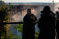 A couple takes photos in Victoria Falls. Behind the bridge between Zambia and Zimbabwe.  The Victoria Falls Bridge crosses the Zambezi River just below the Victoria Falls and is built over the Second Gorge of the falls. As the river is the border between Zimbabwe and Zambia, the bridge links the two countries and has border posts on the approaches to both ends, at the towns of Victoria Falls, Zimbabwe and Livingstone, Zambia. The bridge was the brainchild of Cecil Rhodes, part of his grand and unfulfilled Cape to Cairo railway scheme, even though he never visited the falls and died before construction of the bridge began. Rhodes is recorded as instructing the engineers to "build the bridge across the Zambezi where the trains, as they pass, will catch the spray of the Falls". It was designed by George Anthony Hobson of consultants Sir Douglas Fox and Partners, not as is often stated, Sir Ralph Freeman, the same engineer who contributed to the design of the Sydney Harbour Bridge. At the time of the design of the Victoria Falls Bridge, Freeman was an assistant in the firm who, in those pre-computer days, was calculating stresses. 