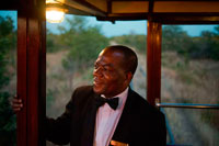 One of the railway workers of the Livingstone Express luxury train. Fine dining is redefined when the venue is the dining car of a 1920’s steam train, in the middle of the Zambian bushveld. The Royal Livingstone Express is a joint venture between Bushtracks Africa and Sun International. This unique dining experience is fully inclusive and guests are transferred from all over Livingstone and the surrounding lodges to the old Mulobezi Line Offices in Livingstone town. Walking up to the train on a red carpet sets the mood for this must-do experience. Each guest is personally greeted and offered a welcome-drink as they board the immaculate vintage first-class Lounge carriage. The Royal Livingstone Express consists of five carriages, restored by Rohan Vos of Rovos Rail and these include two dining cars, a club/kitchen car, lounge car, and an observation car and is pulled by either a 10th class No. 156 or a 12th class No. 204 locomotive. Once all guests are aboard the journey commences through Dambwa suburb towards the Mosi-oa-Tunya National Park, running parallel to the Zambezi River. 