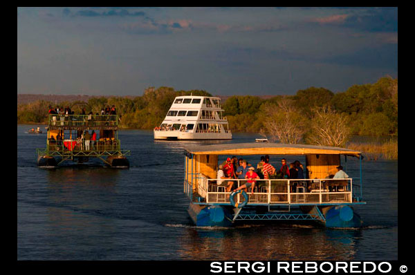 Crucero a lo largo de las Cataratas Victoria a bordo del "African Queen". Otros barcos que navegan en el río Zambezi. Tome un crucero al atardecer por el poderoso río Zambezi. Esta Sundowner crucero te lleva por el río Zambezi para un hermoso viaje!. El crucero Zambezi Puesta del Río es una excelente manera de relajarse y disfrutar de la belleza del río. Usted puede tener la oportunidad de ver una gran variedad de juego, incluyendo hipopótamos, cocodrilos, elefantes e incluso a veces de rinocerontes en su entorno familiar, así como disfrutar de las muchas especies de aves diferentes. El crucero de la puesta del sol es muy popular e incluye aperitivos dedos, cerveza, vino, champán y refrescos. Hay grandes oportunidades de fotos en contra a menudo espectaculares puestas de sol africanas. El crucero tiene una duración de aproximadamente 2 horas.