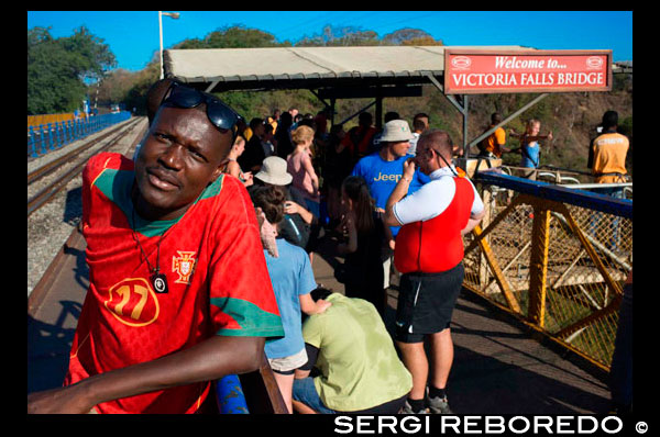 People around Victoria Falls Bridge. The Victoria Falls Bridge crosses the Zambezi River just below the Victoria Falls and is built over the Second Gorge of the falls. As the river is the border between Zimbabwe and Zambia, the bridge links the two countries and has border posts on the approaches to both ends, at the towns of Victoria Falls, Zimbabwe and Livingstone, Zambia. For more than 50 years the bridge was crossed regularly by passenger trains as part of the principal route between the then Northern Rhodesia, southern Africa and Europe. Freight trains carried mainly copper ore (later, copper ingots) and timber out of Zambia, and coal into the country. The age of the bridge and maintenance problems have led to traffic restrictions at times. Trains cross at less than walking pace and trucks were limited to 30 t, necessitating heavier trucks to make a long diversion via the Kazungula Ferry or Chirundu Bridge. The limit was raised after repairs in 2006,but more fundamental rehabilitation or construction of a new bridge has been aired. During the Rhodesian UDI crisis and Bush War the bridge was frequently closed (and regular passenger services have not resumed successfully). In 1975, the bridge was the site of unsuccessful peace talks when the parties met in a train carriage poised above the gorge for nine and a half hours. In 1980 freight and road services resumed and have continued without interruption except for maintenance.
